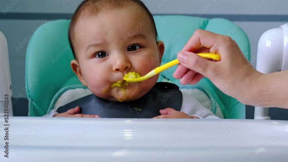 Mom feeds the baby with a spoon of vegetable puree at the children's feeding table. Baby's appetite, healthy nutrition, introduction of complementary foods. Copyspace, mock up Mom feeds the baby with a spoon of vegetable puree at the children's feeding table. Baby's appetite, healthy nutrition, introduction of complementary foods. Copyspace, mock up
