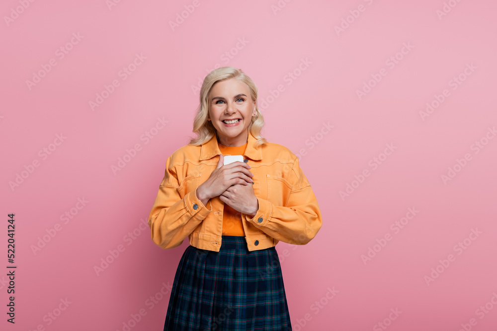 Happy blonde woman holding smartphone and looking at camera isolated on pink Happy blonde woman holding smartphone and looking at camera isolated on pink