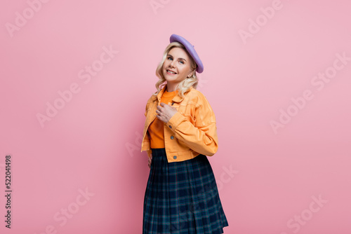 Positive blonde woman in jacket and beret looking at camera isolated on pink