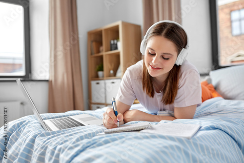 school, online education and e-learning concept - happy smiling teenage student girl in headphones with laptop computer writing to notebook lying on bed at home