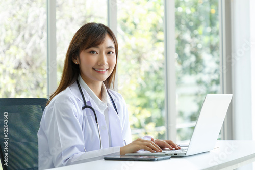 Beautiful and cheerful good mood young lady Asian female doctor sitting in medical office while using laptop computer.
