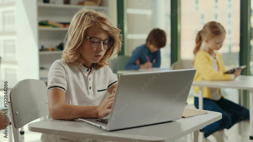 Side view of the red-head boy pupil wearing glasses using laptop for studying purposes at school in the class and other pupils on the background. People and technology concept. Side view of the red-head boy pupil wearing glasses using laptop for studying purposes at school in the class and other pupils on the background. People and technology concept.