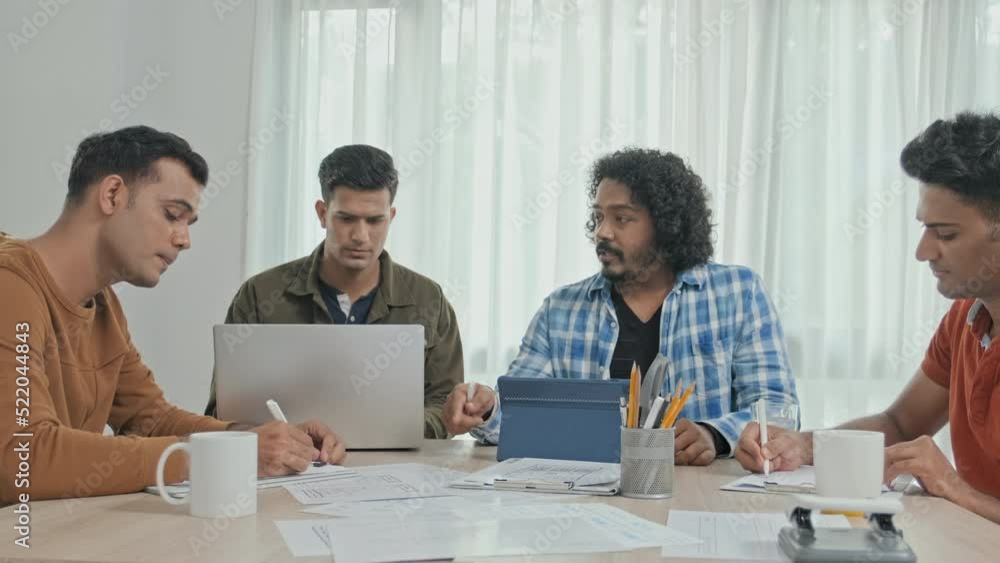 Medium slowmo of four young creative male Indian entrepreneurs discussing business project at table in office Medium slowmo of four young creative male Indian entrepreneurs discussing business project at table in office