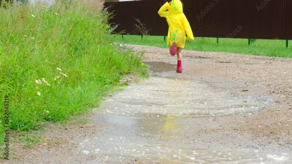 A girl in red rubber boots and a yellow raincoat runs through puddles ...