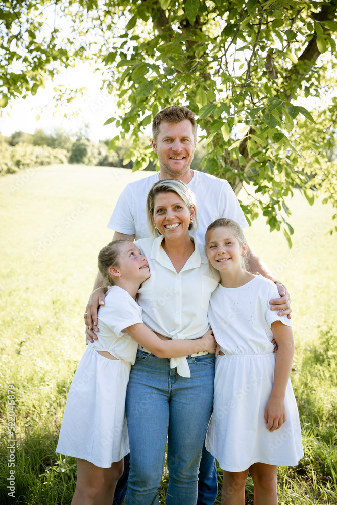 beautiful family with two girls is standing in meadow near walnut tree and has light outfit on and jeans and is happy beautiful family with two girls is standing in meadow near walnut tree and has light outfit on and jeans and is happy