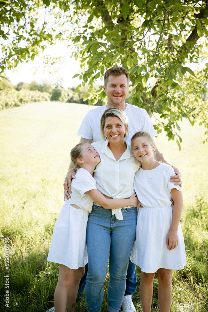 beautiful family with two girls is standing in meadow near walnut tree and has light outfit on and jeans and is happy