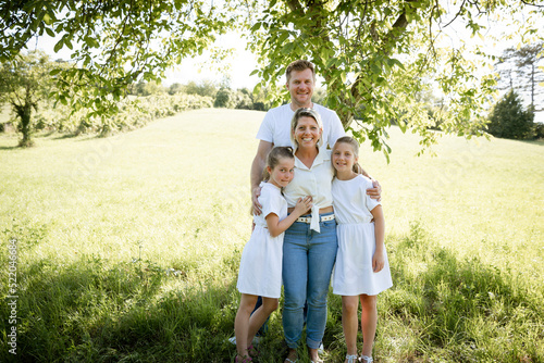 beautiful family with two girls is standing in meadow near walnut tree and has light outfit on and jeans and is happy
