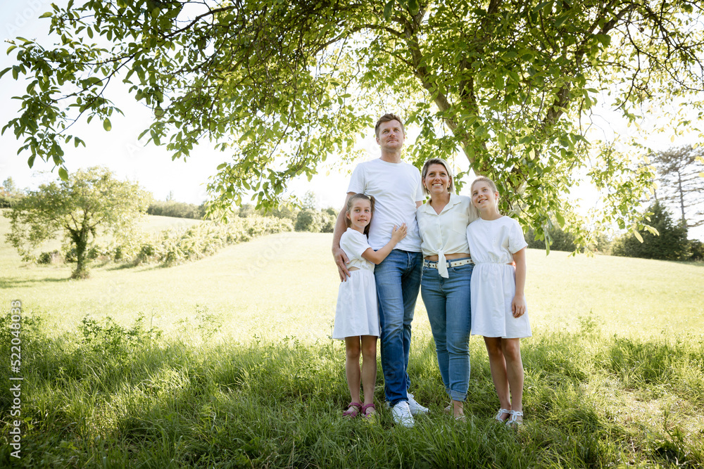 beautiful family with two girls is standing in meadow near walnut tree and has light outfit on and jeans and is happy