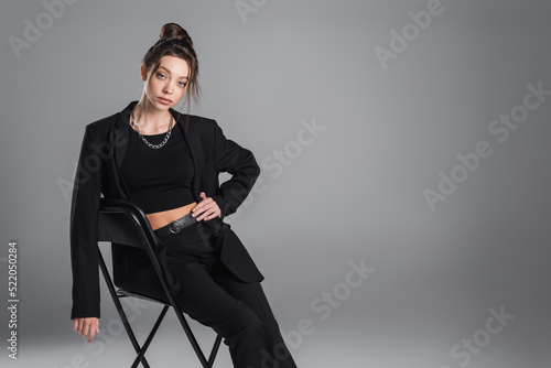 young brunette woman in black trendy suit sitting on chair with hand on hip on grey background
