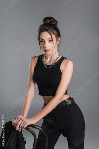 trendy woman in black crop top and silver necklace posing near chair isolated on grey