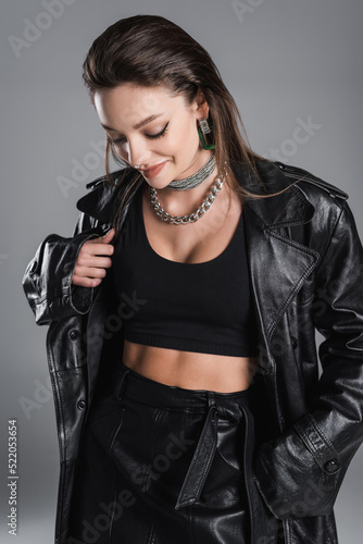 pretty woman in black leather clothes and silver necklaces smiling isolated on grey