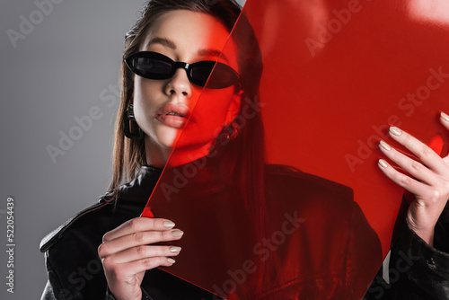young brunette woman in black stylish sunglasses posing with red glass isolated on grey