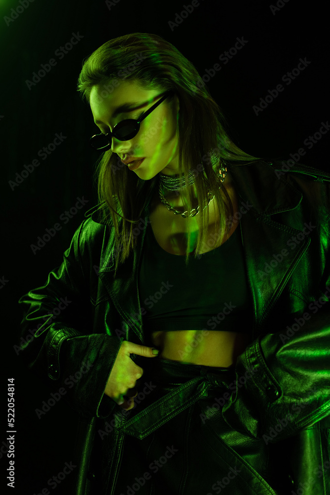 trendy woman in sunglasses and crop top standing with hand on hip in green light isolated on black trendy woman in sunglasses and crop top standing with hand on hip in green light isolated on black