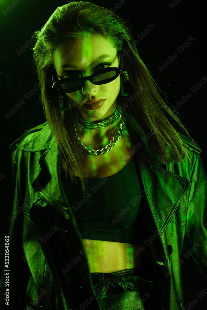 stylish woman in crop top and dark sunglasses looking at camera in green light isolated on black stylish woman in crop top and dark sunglasses looking at camera in green light isolated on black