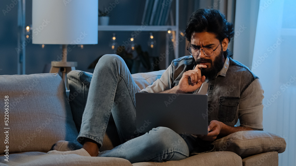 Businessman Arab Indian Hispanic man bearded user wearing glasses sitting on sofa night late time with laptop computer difficulties online trouble thinking idea app pondering solution looking screen Businessman Arab Indian Hispanic man bearded user wearing glasses sitting on sofa night late time with laptop computer difficulties online trouble thinking idea app pondering solution looking screen