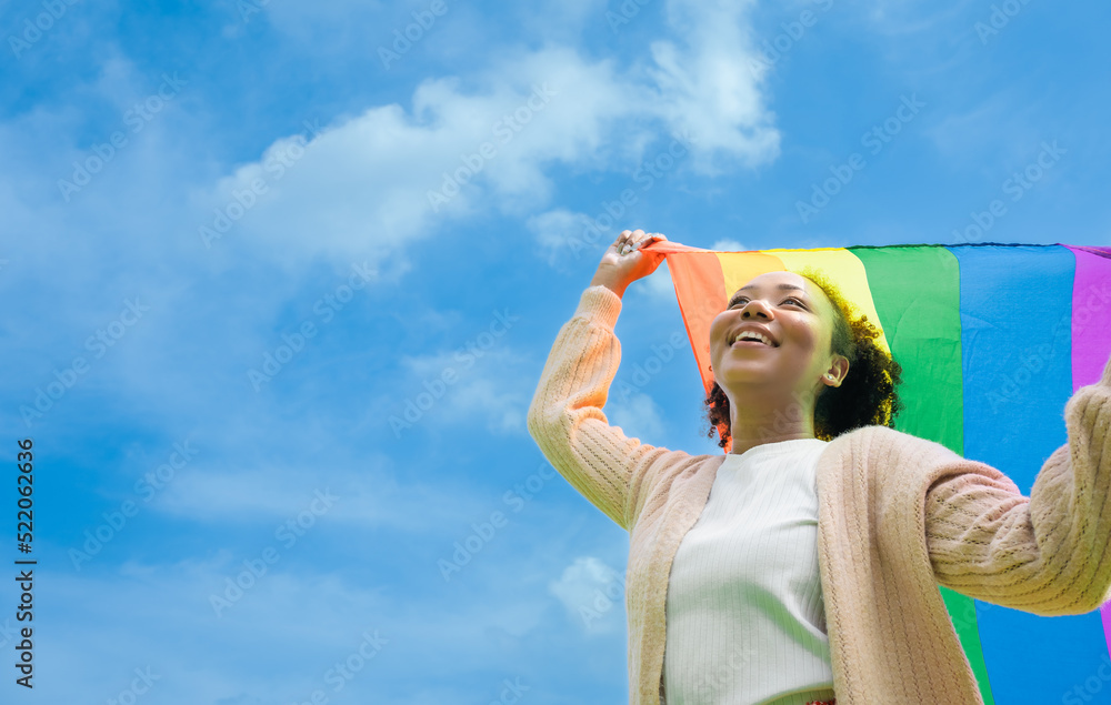 Naklejka premium lgbtq concept. Positive African girl with afro curly hair holding rainbow flag isolated in studio.An African American young woman holding and raising a rainbow flag over the blue sky.
