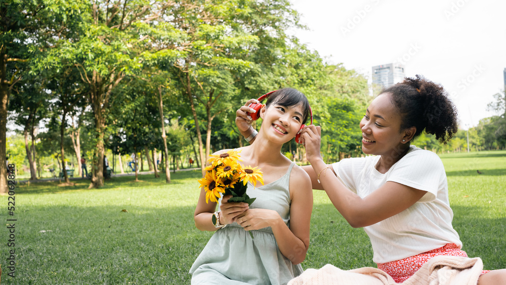 Fototapeta premium Smiling two young diversity African American Asian women friends having fun in a nature, park queer non-binary gender identity, gay lesbian love romance, boho summer vacation style.Listening music.