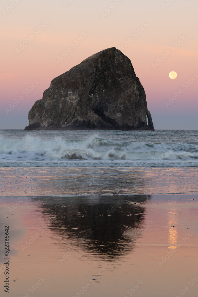 Waves crash ashore infront of Haystack Rock at Pacific City Oregon at dawn as the full moon sets on a colorful horizon.  The monolith reflects in the wet beach.