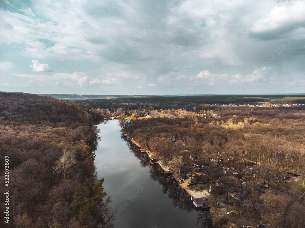 Aerial view on river curve in bare trees forest with epic cloudy sky. Nature landscape from drone near Koropove village in Kharkiv region. Color graded