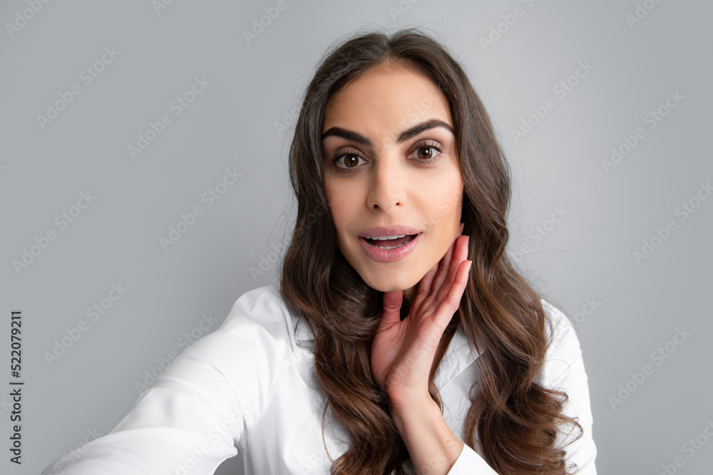 Portrait of cheerful smiling woman having video-call, shooting selfie. Young girl in white shirt with dark long hair isolated on gray background taking selfie photo, posing, smiling playful. Portrait of cheerful smiling woman having video-call, shooting selfie. Young girl in white shirt with dark long hair isolated on gray background taking selfie photo, posing, smiling playful.
