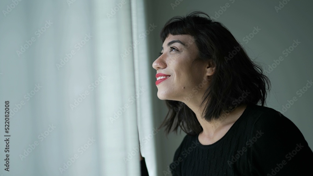 One happy hispanic woman standing by window smiling looking out seeing beautiful day One happy hispanic woman standing by window smiling looking out seeing beautiful day