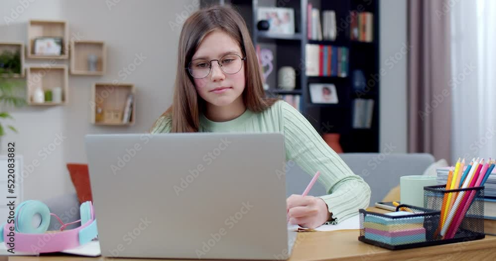 Pretty smart schoolgirl in glasses sitting at laptop, writing exercise at home as having online class. Schooling by Internet. Teenage girl learning at computer at home, write task. Education concept. Pretty smart schoolgirl in glasses sitting at laptop, writing exercise at home as having online class. Schooling by Internet. Teenage girl learning at computer at home, write task. Education concept.