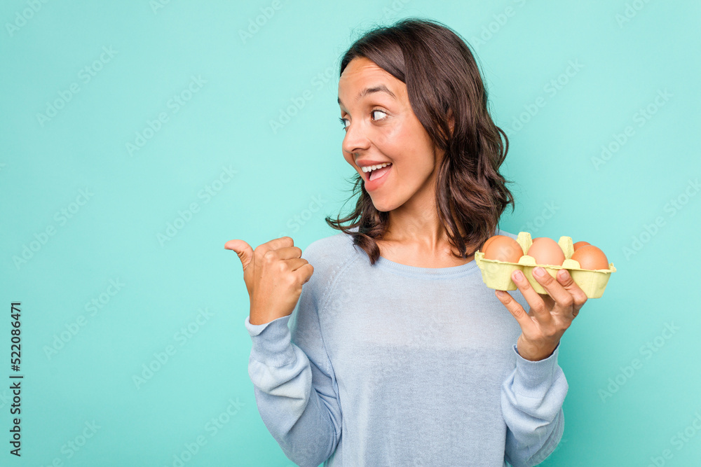 Young hispanic woman holding eggs isolated on blue background points with thumb finger away, laughing and carefree. Young hispanic woman holding eggs isolated on blue background points with thumb finger away, laughing and carefree.