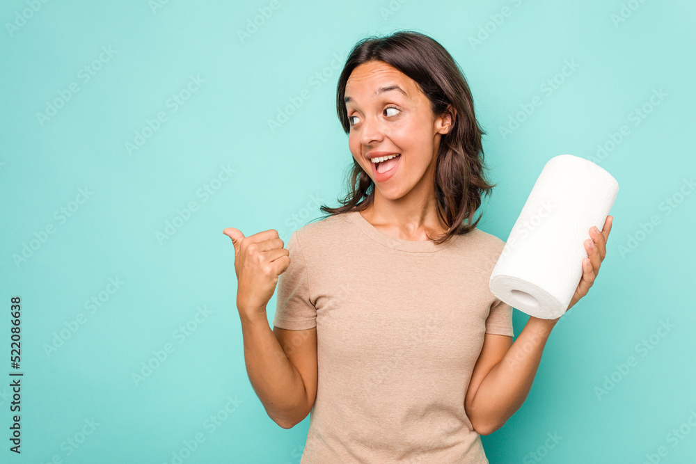 Young hispanic woman holding kitchen roll isolated on blue background points with thumb finger away, laughing and carefree. Young hispanic woman holding kitchen roll isolated on blue background points with thumb finger away, laughing and carefree.
