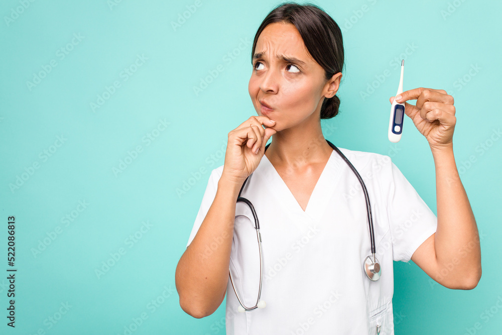 Young hispanic nurse woman holding a thermometer isolated on blue background looking sideways with doubtful and skeptical expression. Young hispanic nurse woman holding a thermometer isolated on blue background looking sideways with doubtful and skeptical expression.
