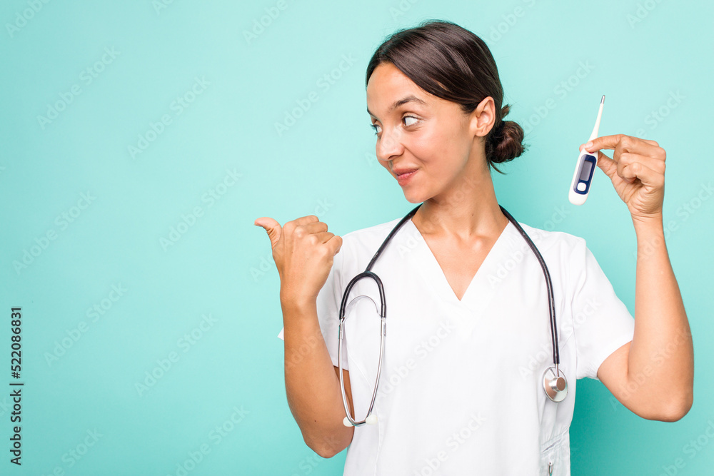 Young hispanic nurse woman holding a thermometer isolated on blue background points with thumb finger away, laughing and carefree. Young hispanic nurse woman holding a thermometer isolated on blue background points with thumb finger away, laughing and carefree.