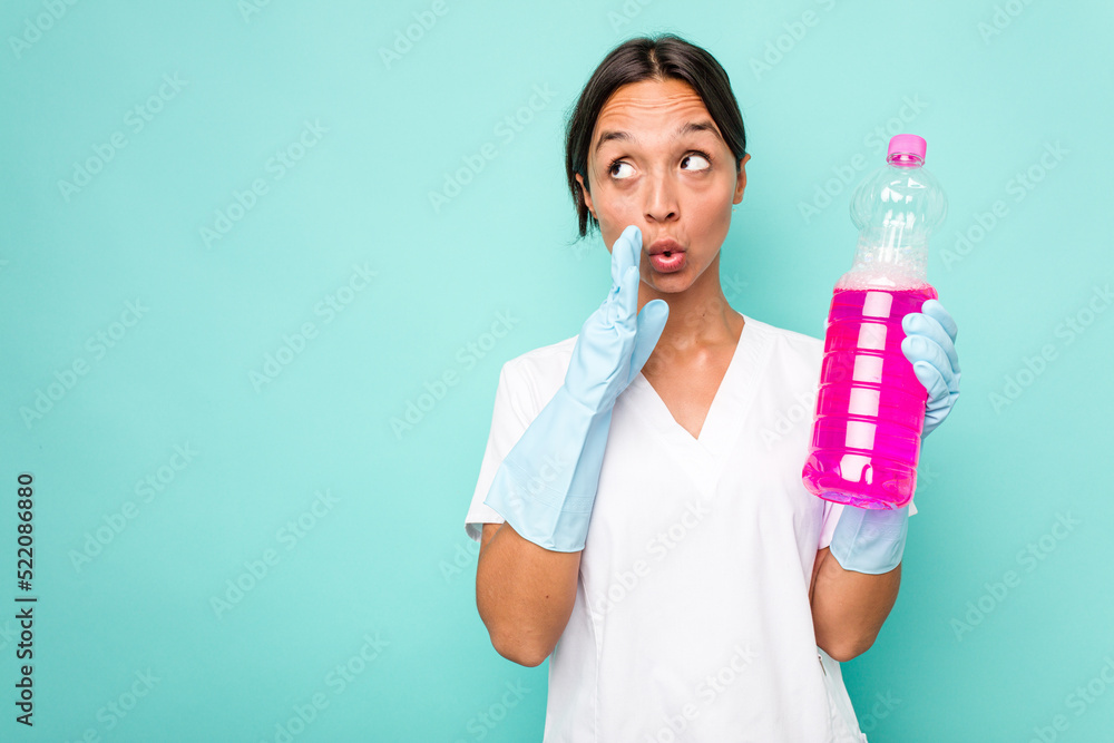 Young cleaner hispanic woman isolated on blue background is saying a secret hot braking news and looking aside Young cleaner hispanic woman isolated on blue background is saying a secret hot braking news and looking aside