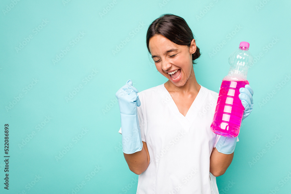 Young cleaner hispanic woman isolated on blue background raising fist after a victory, winner concept. Young cleaner hispanic woman isolated on blue background raising fist after a victory, winner concept.