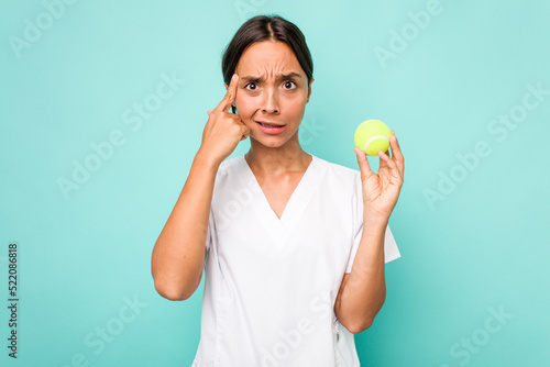 Young hispanic physiotherapy holding a tennis ball isolated on blue background showing a disappointment gesture with forefinger. © Asier