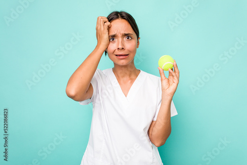 Young hispanic physiotherapy holding a tennis ball isolated on blue background being shocked, she has remembered important meeting. © Asier
