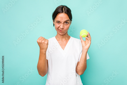 Young hispanic physiotherapy holding a tennis ball isolated on blue background showing fist to camera, aggressive facial expression. © Asier