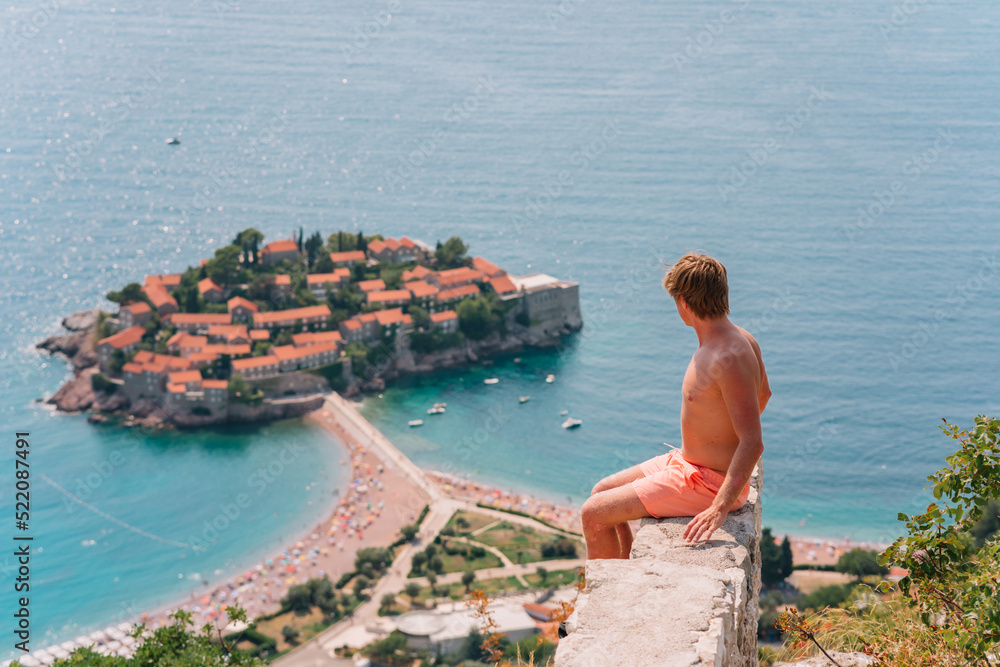 Obraz premium a man poses on a mountain with a view from above of the city of Sveti Stefan in Montenegro, a picturesque bay