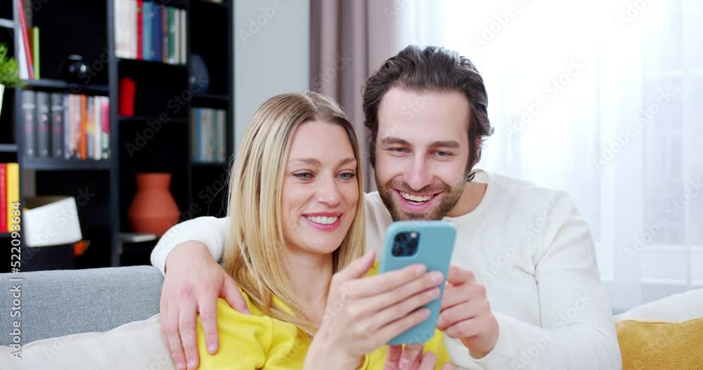 Joyful Caucasian wife and husband sitting on sofa in cozy room and tapping on mobile phone. Indoors. Beautiful woman and handsome man scrolling on mobile phone in hugs at home. People with gadget.