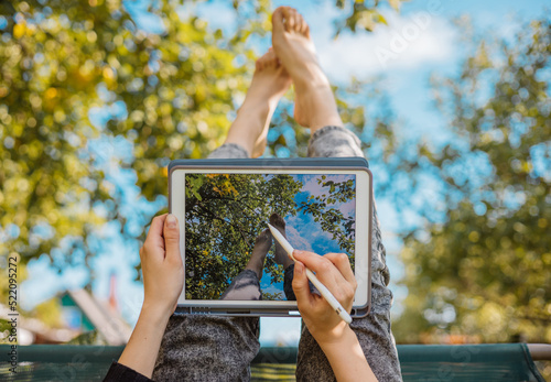 Teenage girl sits in the garden on a swing upside down and draws on a tablet sketches with her legs on background of the sky. Inspiration, creativity, self-development, design. Modern technologies