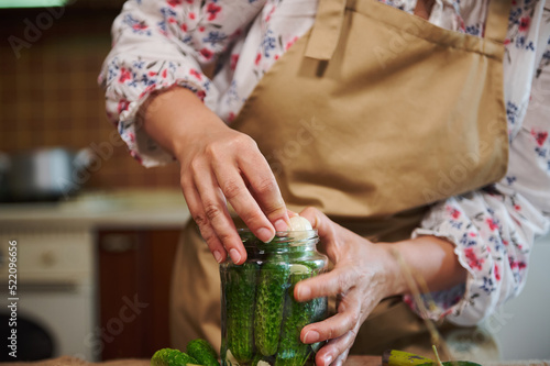 Details: Hands of a woman housewife adding fresh garlic clove in a glass jar while preparing homemade pickled cucumbers. Preservation of fresh house cucumbers. Selective focus.