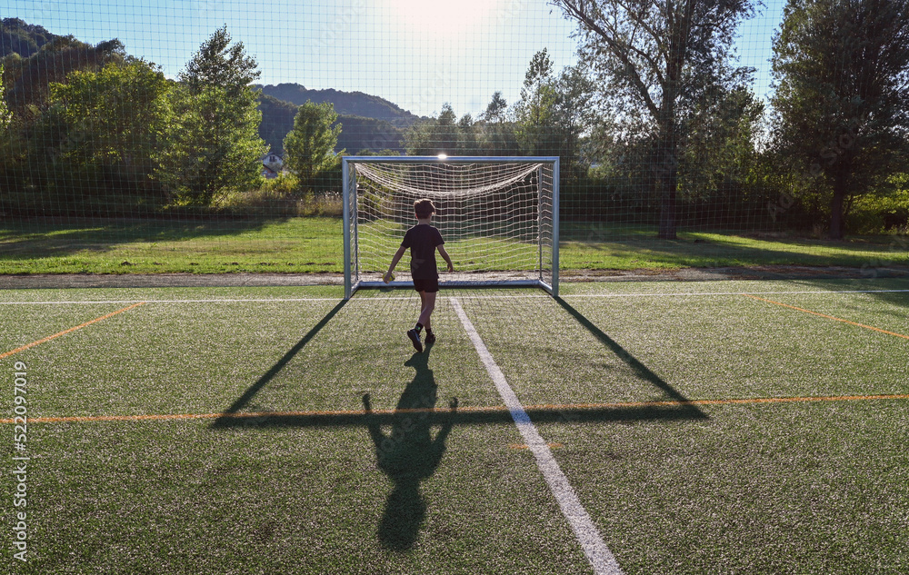 Fototapeta premium Young boy playing soccer on the football field at sunset, shooting at empty goal posts