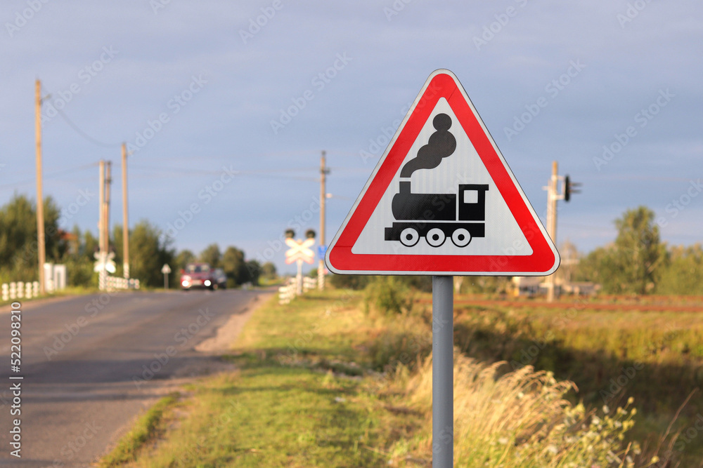Railway road sign in red and white, railway crossing sign, in the background a car is moving over the rails Railway road sign in red and white, railway crossing sign, in the background a car is moving over the rails