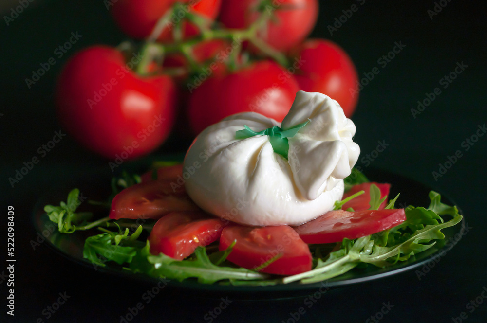 on a black plate, spinach salad and arugula greens, sliced tomatoes and Italian burrata cheese on a black background, a bunch of red tomatoes is visible in the background, side view. on a black plate, spinach salad and arugula greens, sliced tomatoes and Italian burrata cheese on a black background, a bunch of red tomatoes is visible in the background, side view.