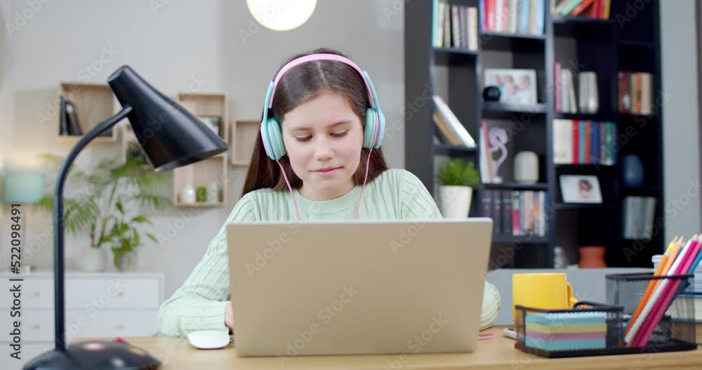 Caucasian teen pretty smiled girl in headphones playing on laptop and listening to music at home. Beautiful happy kid chatting online with smile in cozy living room and enjoying song. Inside. Caucasian teen pretty smiled girl in headphones playing on laptop and listening to music at home. Beautiful happy kid chatting online with smile in cozy living room and enjoying song. Inside.