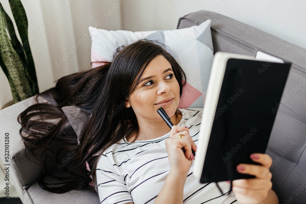 Young Brazilian woman lying on the sofa writing a diary. Woman writing notes at home. Young Brazilian woman lying on the sofa writing a diary. Woman writing notes at home.