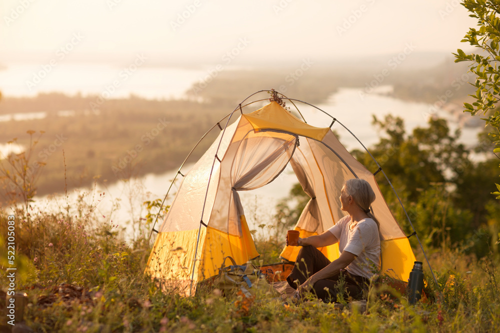portrait of a smiling senior woman relaxed in a tent and drinks tea outdoors at sunset. copy space. Slow life. Enjoying the little things. spends time in nature in summer