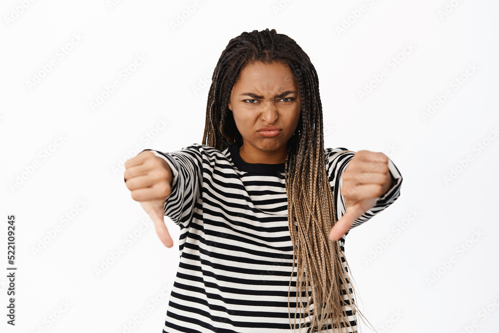 Portrait of disappointed black girl grimacing, showing thumbs down to express dislike and aversion, do not recommend smth, white background Portrait of disappointed black girl grimacing, showing thumbs down to express dislike and aversion, do not recommend smth, white background