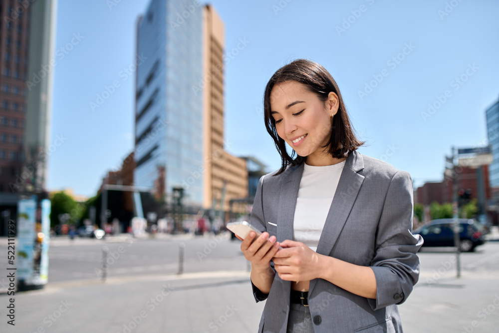 Young adult smiling professional Asian business woman wearing suit holding smartphone looking at mobile phone using apps on cellphone technology texting standing on urban city street outside.