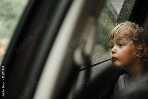 Thoughtful little blond baby boy seated in car backseat looking through rain covered window in rainy day.