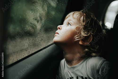 Thoughtful little blond baby boy seated in car backseat looking through rain covered window in rainy day.