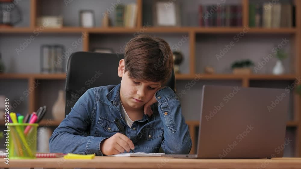 Young boy studying remotely at home using laptop. Sad schoolboy feeling bored doing homework or during an online lesson with teacher. Distance learning and home education. Young boy studying remotely at home using laptop. Sad schoolboy feeling bored doing homework or during an online lesson with teacher. Distance learning and home education.
